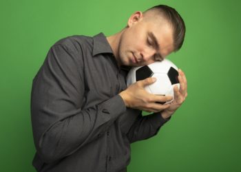 Homem em pé vestido de camisa social cinza chumbo, com uma bola na mão e nela a cabeça de olhos fechados, como estivesse dormindo